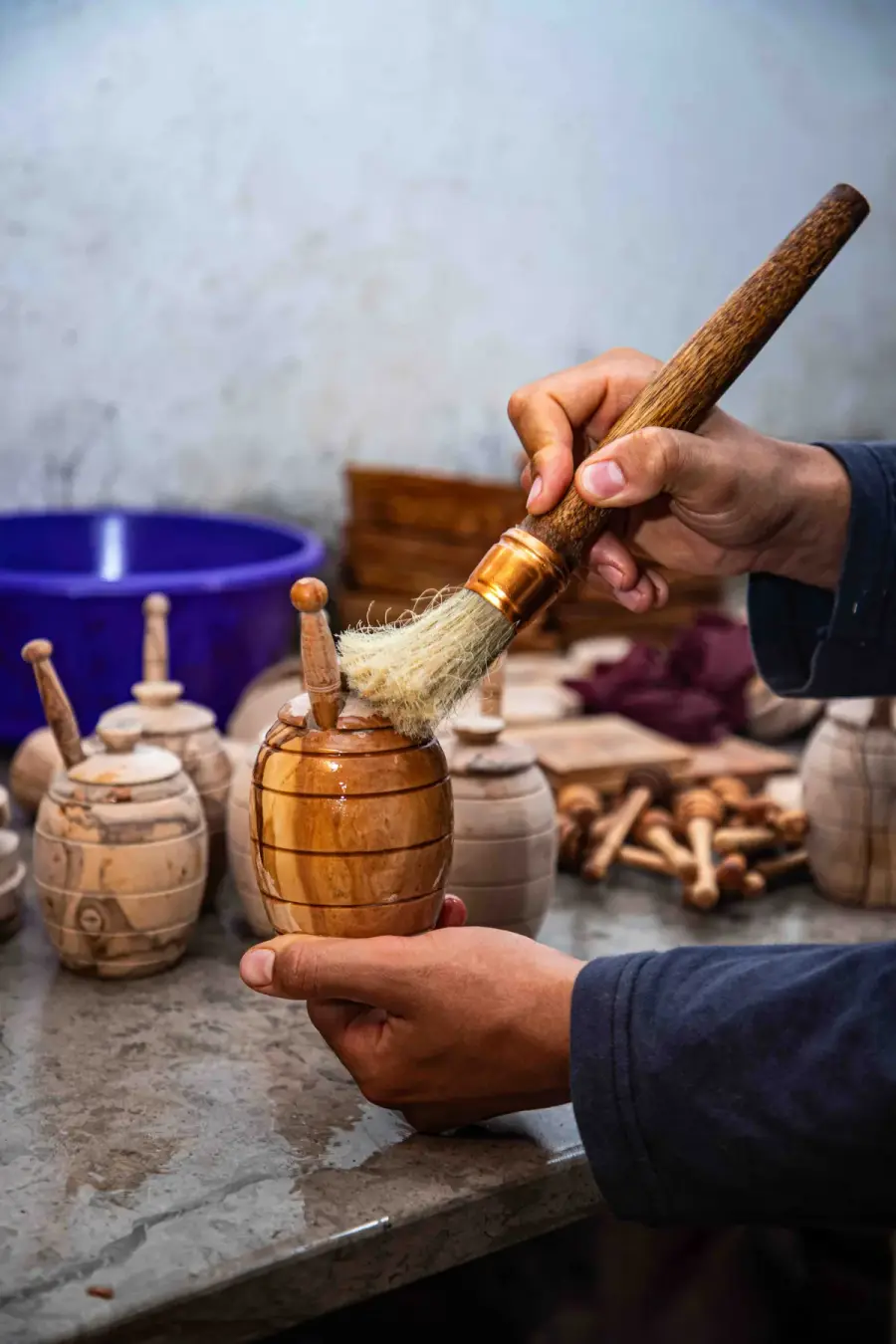 A worker brushing oil onto a small turned olive wood jar, with unfinished jars and pestle handles waiting on the bench